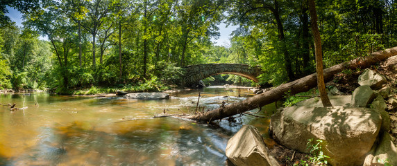 A panoramic shot of Boulder Bridge in Rock Creek Park in Washington, DC on a summer day. Rock Creek flows underneath the iconic bridge; a fallen tree is seen in the foreground.