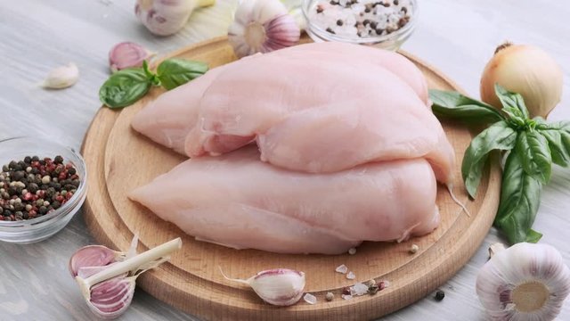 Raw chicken fillet on a round wooden board. Heads of garlic, basil, pepper, salt. Culinary concept. Slow rotation. 4k
