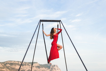 Woman in red dress practicing pole fly dance poses in a hammock outdoor with a mountain view. Female sports, wellbeing concept. Pilates outdoors.