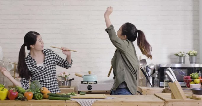 Happy Young Asian Girls Dancing To Music Together Enjoying Cooking In Kitchen. Two Woman Having Fun Preparing Breakfast. Chinese Female Friends Singing With Spatulas As Microphone At Home In Day Time