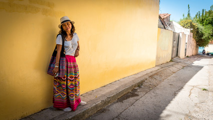 Asian woman in white t-shirt  in Athens in white hat and sarong leaning on yellow wall