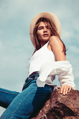 Beautiful girl in a white shirt, blue jeans and cowboy hat posing on the rock. Outdoor shot. Summer vacation concept.