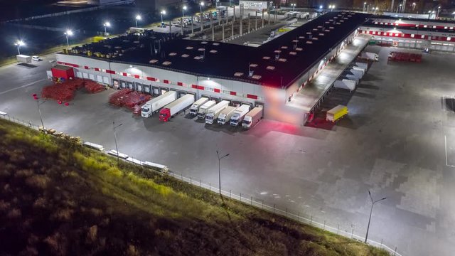Aerial hyper lapse (motion time lapse), view of the large logistics park with warehouse, loading hub with many semi-trailers trucks standing at the ramps for load/unload goods at night