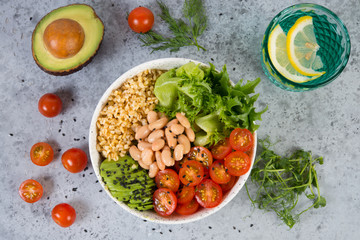 A plate of fresh salad with white beans, bulgur, cherry tomatoes and avocado, decorated with black sesame seeds with products around the plate. Horizontal photo with copy space, top view