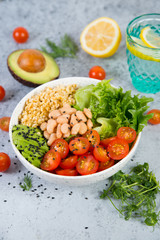 A plate of fresh salad with white beans, bulgur, cherry tomatoes and avocado, decorated with black sesame seeds with products around the plate.