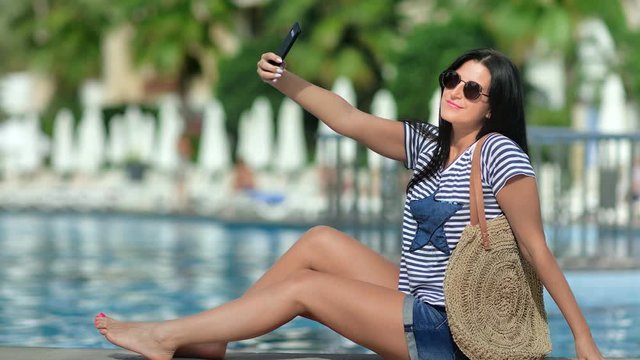 Smiling travel woman taking selfie using smartphone at swimming pool palm background