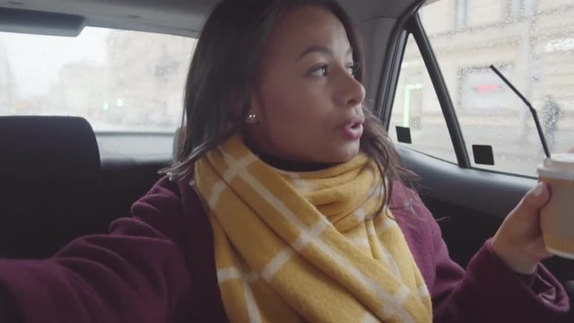 Chest-up handheld shot of happy young mixed race woman in coat and scarf, holding takeaway coffee cup and invisible smartphone in outstretched hand, riding in back of moving taxi and video chatting