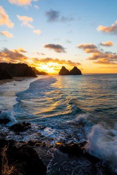 Stunning sunset in Fernando de Noronha, Brazil. Taken from a mountain peak called Pedra do Bode. Two sets of waves crashing at rocks with sun setting at distance near Two Brothers Mountain.