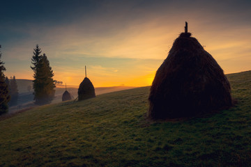 Beautiful sunrise over mountain foggy hills. Scenic landscape with sun, rising over Carpathian mountains. Haystack on grassland hill on foreground.