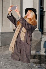 Naklejka premium A young woman talks on her smartphone and takes a selfie. A teenager in a gray coat on the street. Winter fashion.