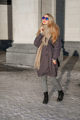 Fototapeta premium A beautiful woman in a gray coat and sunglasses walks on the street in the city.