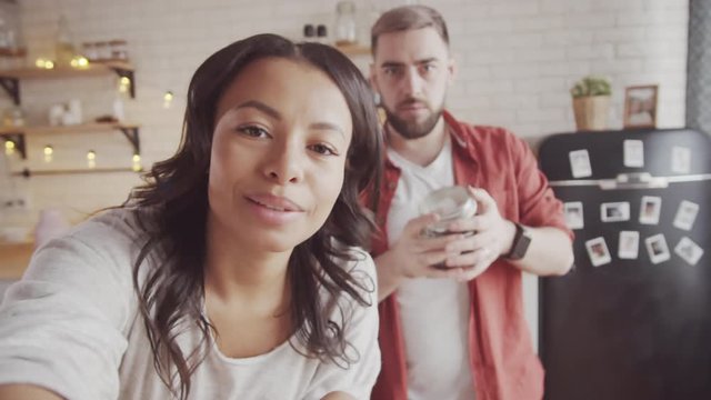 Medium handheld shot of happy married couple video calling someone in kitchen decorated with Christmas lights at home, looking at camera, smiling and showing mug and jar of coffee beans