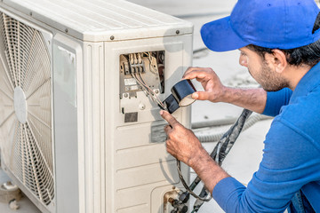 a professional electrician man is fixing a heavy duty unit of central air conditioning system by his tools on the roof top and wearing blue color of uniform and white cap