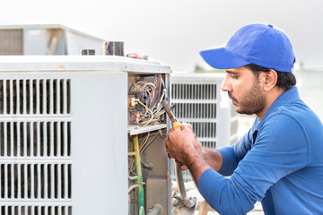 a professional electrician man is fixing the heavy unit of an air conditioner at the roof top of a building and wearing blue uniform and head cap