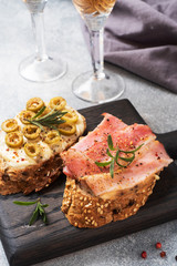 Wholegrain bread sandwiches with cream cheese, bacon and olives on a wooden cutting Board. A glass of white wine. Gray concrete background.