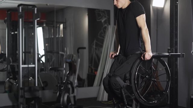 Workout, young disabled athlete in wheelchair performs push-ups on bars, gym training and physical activity.