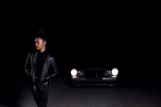 A Young  Mulatto Girl In A Leather Jacket And Black Clothes Poses On An Abandoned Sand Road. At Night In The Light Of Car Headlights And Lanterns