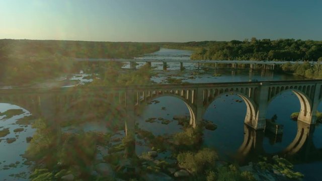 Medium wide sunny aerial drone shot pushes along the James River toward, and tilting down onto, the ACL Railroad Bridge and the Powhite Parkway outside Richmond Virginia