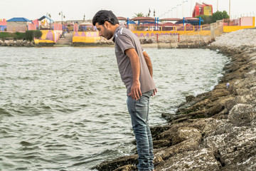 a lonely man is standing on the sea rocks and thinking for suicide after divorce and wearing grey t shirt and blue jeans in the bright cloudy day