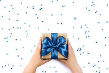 Child's hands holding present with blue bow on white rustic sparkling background. Flat lay style.
