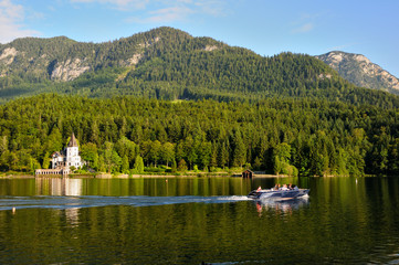 A large white house in the form of a castle on the shore of the lake. Green mountains in the background. Motor boat on the lake.