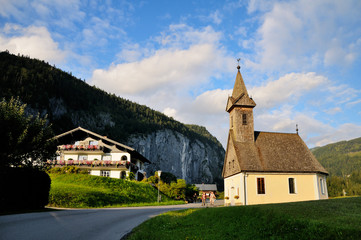 Fototapeta na wymiar A small Catholic church with a wooden roof in a Bavarian village. Beautiful evening lighting. Blue sky and mountains in the background.