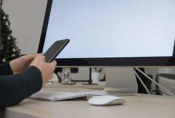 Obraz premium Women's hands hold the phone in front of the computer. Focus on computer screen. Christmas trees in the back of the office. The woman works during the Christmas and New Year holidays.