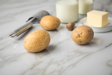 Fototapeta na wymiar Kartoffeln, Milch, Salz, Butter, Muskatnuss als Zutaten mit Kartoffelpresse und Muskatreibe für Kartoffelpüree auf hell Marmor Hintergrund