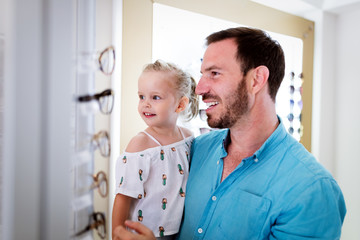 Obraz premium Health care, eyesight and vision concept. Little girl choosing glasses with father at optics store