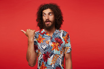 Fototapeta na wymiar Studio photo of young handsome brunette curly male with beard looking scaredly aside with wide eyes opened, pointing aside with thumb while posing over red background