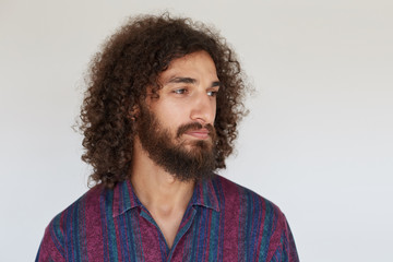 Fototapeta na wymiar Portrait of pretty young brunette curly man with beard keeping lips folded while looking aside with calm face, wearing striped multi-colored shirt while posing against white background