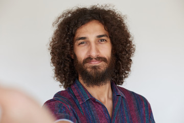 Attractive positive brunette curly man with beard holding camera and smiling sincerely while making...