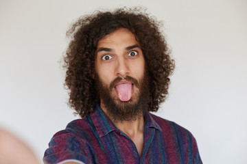 Funny indoor shot of attractive young dark haired curly bearded man fooling and making faces while standing against white background, dressed in striped multi-colored shirt