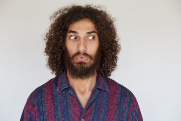 Fototapeta na wymiar Indoor photo of bemused young brunette curly bearded male dressed in casual clothes folding lips and rounding eyes confusedly while posing against white background