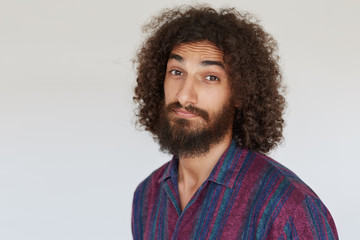 Fototapeta na wymiar Close-up of handsome young bearded brown-eyed man with dark curly hair raising eyebrows and wrinkling forehead while looking at camera, isolated against white background