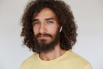Close-up of beautiful young dark haired curly man with charming brown eyes looking positively to camera with light smile, wearing lush beard and listening to music in his headphones