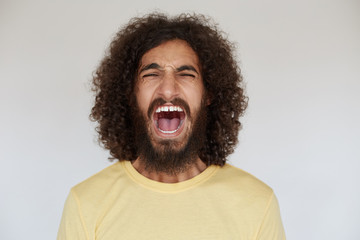 Studio shot of stressed young dark haired curly male with beard screaming loud with wide mouth opened and frowning his face, keeping hands down while posing over white background