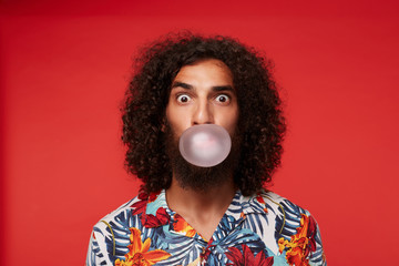 Amazed pretty brown-eyed bearded guy with dark curly hair making bubble with gum and looking...