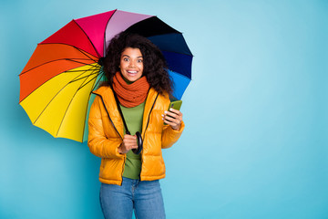 Photo of pretty dark skin lady hold colorful umbrella telephone modern social network blogger wear stylish overcoat jeans green pullover scarf isolated blue color background © deagreez