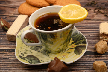 A Cup of black tea, tea leaves, pieces of brown sugar, oatmeal cookies, waffles, pieces of chocolate, lemon on a wooden background. Close up.