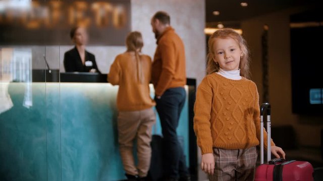 A family checking in the hotel - a little girl standing with a suitcase and waiting for her parents