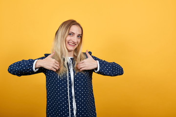 Smiling young woman in blue shirt with white polka dot over isolated orange background, pointing with thumbs at herself. Lifestyle concept