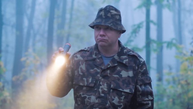 A man in camouflage is walking through the forest with a flashlight in his hand. Wet rainy weather in the early morning