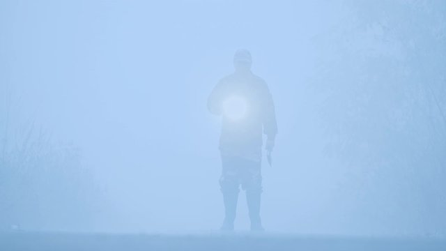 ilhouette of a man with a flashlight and an ax walking in the fog