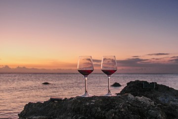 Two glasses of red wine  on the  rock beach near water. Romantic date on a beautiful sunset ocean background.  Romantic glasses of red wine against a colorful sunset.