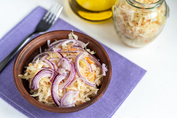 Fermented, pickled cabbage in a brown plate with red onion on a gray background. Behind the jar and a sauce boat with vegetable oil. Copy space.