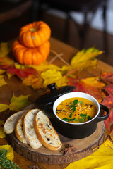 Warm and spicy carrot soup served with bread and cardamon on a table in black pot with lid. Surrounded by yellow and read leaves, pumpkins on background