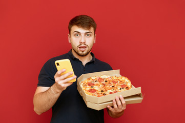 Man with a beard stands on a red background with an open pizza box and a smartphone in his hand, looking into the camera with a surprised face. Man received a pizza ordered online.