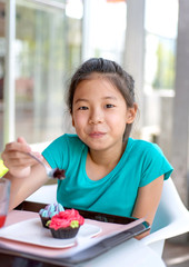Smiling Asian preteens girl enjoy eating tasty cup cake with whipping cream in dining room. Cute teenage girl holding spoon with sweet dessert looking at camera.