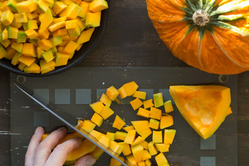 Cutting piece of pumpkin with kitchen knife by woman hand on glass cutting board on wooden background